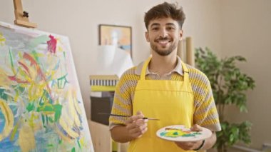 Gülümseyen Arap genç adam resim atölyesinde boya fırçası yeteneğini sergiliyor, resim hobisini ve eğitimini özgüvenle kucaklıyor.