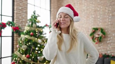 Genç sarışın kadın Noel 'i kutluyor ve evde akıllı telefondan konuşuyor.