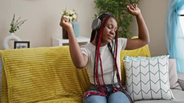 Güzel örgüleri olan neşeli Afrikalı Amerikalı kadın evdeki kanepesinde dans ediyor kulaklıklarıyla dinlediği müziğe ayak uyduruyor. Kendi eğlence dolu ev yaşamının tadını çıkarıyor..