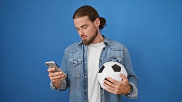 Young hispanic man holding soccer ball and smartphone looking upset over isolated blue background