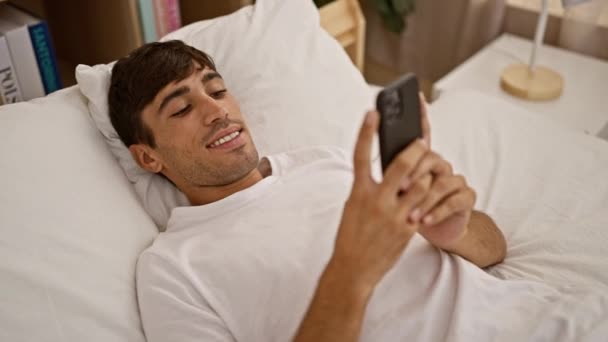 快適にベッドに横たわり 居心地の良い寝室の家のインテリアで輝く笑顔でスマートフォンにテキストを送る魅力的な若いヒスパニック男性 — ストック動画