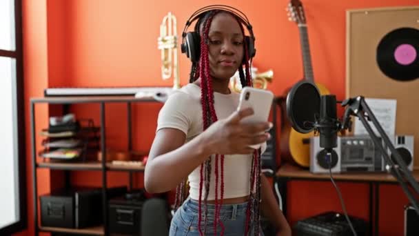 微笑着非洲裔美国女音乐家在一个充满活力的音乐工作室里用她的智能手机 带着甜蜜的自拍 带着耳机 — 图库视频影像