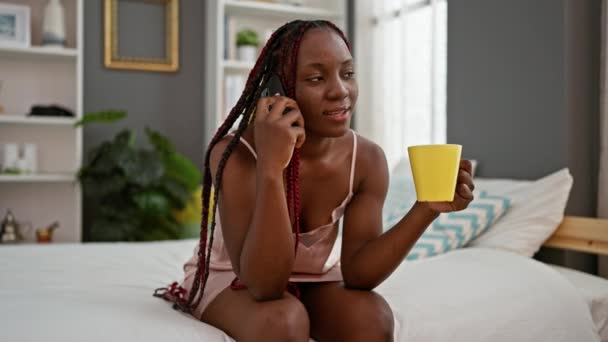 自信的非洲裔美国女人躺在床上休息 在舒适的卧室里 一边用智能手机一边喝咖啡一边愉快地聊天 — 图库视频影像