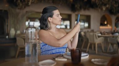 Genç İspanyol kadın restoranda masada oturmuş akıllı telefonuyla fotoğraf çekiyor.