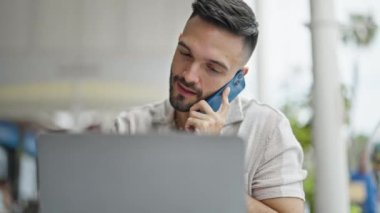 Latin kökenli genç adam, kafenin terasında bilgisayarla akıllı telefondan konuşuyor.
