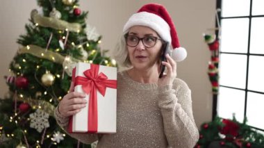 Beyaz saçlı, akıllı telefonlu orta yaşlı bir kadın Noel ağacının yanında duruyor.