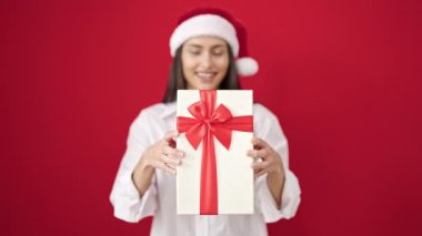 Genç, güzel, İspanyol bir kadın, gülümsüyor. Kendine güveniyor, kırmızı arka planda bir Noel hediyesi tutuyor.