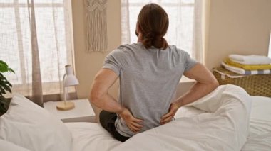 Young hispanic man suffering for backache sitting on bed at bedroom