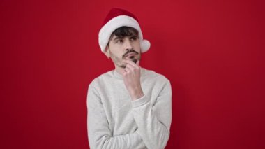 Noel şapkası takmış gülümseyen genç İspanyol adam izole edilmiş kırmızı arka planda bir fikir ediniyor.