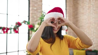 Genç ve güzel İspanyol kadın Noel ağacının yanında gülümsüyor. Elleriyle kalp şekli yapıyor.