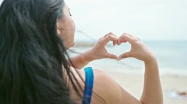 Genç Çinli kadın turist bikini giyip plajda kalp hareketi yapıyor.