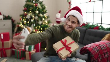 Genç İspanyol adam kamerayla selfie çekiyor. Evdeki Noel ağacının yanında, koltukta oturuyor.