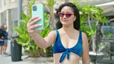 Bikini giyen genç Çinli kadın turist parkta akıllı telefondan selfie çekiyor.