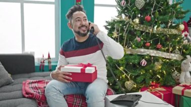 Genç latin adam telefonda konuşuyor. Evde Noel hediyesi tutuyor.