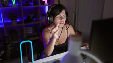 Aşırı çalışılmış ve stresli genç bir İspanyol kadın, oyun dünyasının karanlık yüzünü gece oyun odasında, dijital stres içinde endişeyle oynarken gösteriyor..