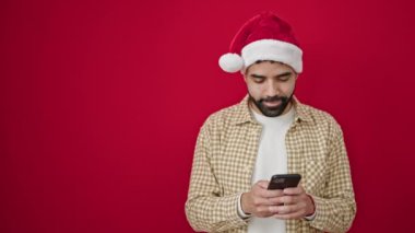 Genç İspanyol adam yılbaşı şapkası takıyor ve izole edilmiş kırmızı arka plan üzerinde akıllı telefon kullanıyor.