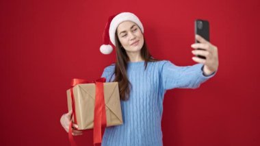 Kafkasyalı genç bir kadın noel şapkası takıyor ve izole edilmiş kırmızı arka planda akıllı telefon hediyesiyle selfie çekiyor