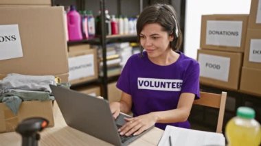Neşeli genç İspanyol kadın gönüllü, neşeli bir şekilde gülümsüyor, hayırseverlik merkezinde özverili online hizmetini kutluyor, bilgisayarında kulaklıklarla ev içi sıcak bir ortamda çalışıyor.