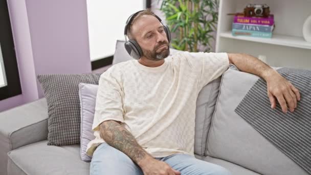 Yakışıklı Genç Adam Oturma Odasındaki Kanepede Rahatça Oturuyor Kulaklıklarından Dökülen — Stok video