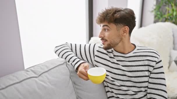 年轻英俊的阿拉伯男子流露出自信 一边享受着早晨的咖啡 一边快乐地微笑着 一边舒服地坐在家里的沙发上 — 图库视频影像