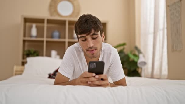 ビクトリア朝 魅力的な若いヒスパニック男性は快適にベッドに横たわり 居心地の良い寝室でスマートフォンを使用して 広い笑顔で勝利を祝います — ストック動画