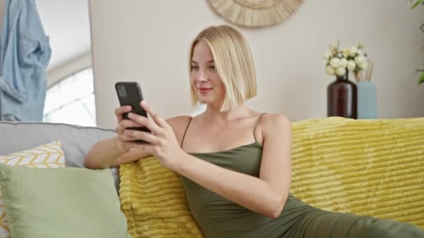 若いですブロンド女性使用してスマートフォン座っています上のソファにホーム — ストック動画