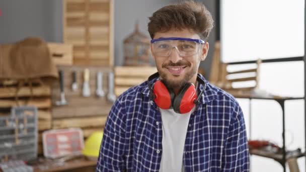 英俊的年轻的阿拉伯男子 戴着眼镜的喜形于色的木匠 兴高采烈地在他那熙攘的室内木工作坊里炫耀着一个大拇指 — 图库视频影像