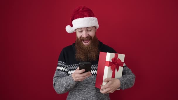Jonge Roodharige Man Met Kerstcadeau Met Behulp Van Smartphone Geïsoleerde — Stockvideo