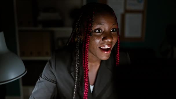 有自信的非洲裔美国女性老板头戴辫子庆祝自己的商业成功 同时还在一个明亮的夜晚在漂亮的办公室里的电脑上工作 — 图库视频影像
