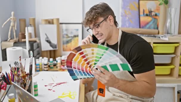 穿着围裙 俊俏的年轻男艺人在智能手机上聊天 为画布选用迷人的色彩 沉浸在繁华的艺术工作室里的创意对话中 — 图库视频影像
