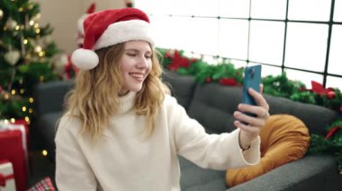 Young blonde woman having video call sitting on sofa by christmas tree at home