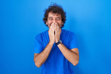 Mavi arka planda dikilen İspanyol genç adam gülüyor ve utanıyor kıkır kıkır kıkır kıkır gülüyor elleri, dedikoduları ve skandalları anlatıyor 