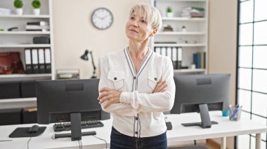 Orta yaşlı, sarışın, iş kadını gülümseyen, kendine güvenen, kollarını kavuşturup ofiste el kol hareketi yapan bir kadın.