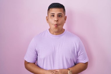 Young hispanic man standing over pink background making fish face with lips, crazy and comical gesture. funny expression. 