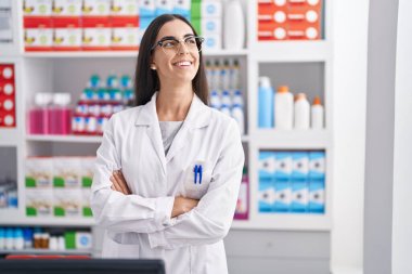 Young beautiful hispanic woman pharmacist smiling confident standing with arms crossed gesture at pharmacy