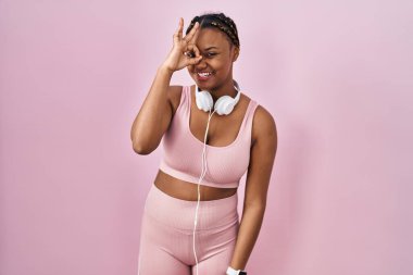 Spor kıyafetleri ve kulaklıkları olan Afro-Amerikalı bir kadın el hareketleriyle gülümsüyor ve mutlu yüzüyle parmaklarının arasından bakıyor.. 