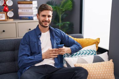 Beyaz bir adam akıllı telefon kullanıyor. Evdeki kanepede oturuyor.