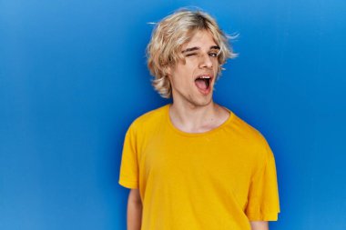 Genç modern bir adam mavi arka planda duruyor. Seksi bir ifadeyle kameraya bakıyor. Neşeli ve mutlu bir yüzü var.. 