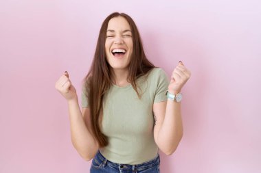 Güzel esmer kadın pembe arka planda mutlu ve heyecanlı bir şekilde kolları havada, gülümseyen ve başarı için bağıran kazanan bir jest yapıyor. kutlama konsepti. 