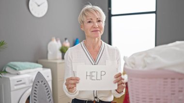 Orta yaşlı sarışın kadın çamaşırhanede çamaşır ipine kıyafet asmak için yardım istiyor.