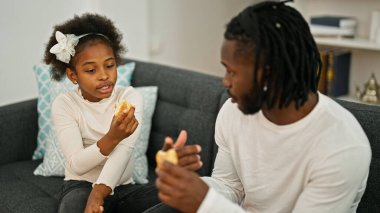 Afrikalı Amerikalı baba ve kız kanepede oturmuş evde kruvasan yiyorlar.
