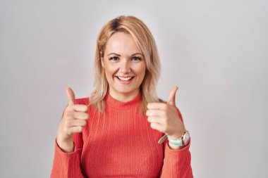 Sarışın kadın arka planda izole edilmiş başarı işaretinin başında duruyor. Elleriyle olumlu jest yapıyor. Gülümsüyor ve mutlu. Neşeli ifade ve kazanan jest. 