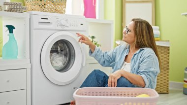 Orta yaşlı İspanyol kadın çamaşırhanede yerde oturmuş çamaşır yıkıyor.