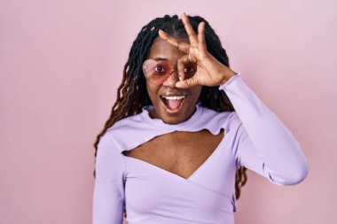 Pembe arka planda örülü saçları olan Afrikalı kadın şaşırmış yüz ifadesiyle şaşırmış, parmaklarının arasından bakan bir jest yapıyor. İnançsız ifade. 