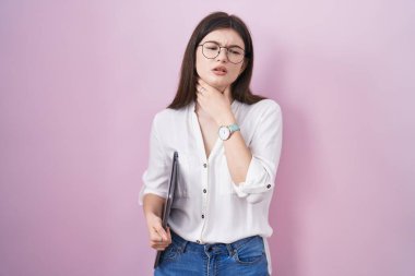 Genç, beyaz bir kadın dizüstü bilgisayar tutuyor. Boynuna dokunuyor. Grip, mantar ve enfeksiyon yüzünden boğaz ağrısı çekiyor. 