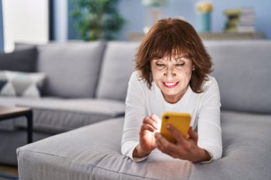 Middle age woman using smartphone lying on sofa at home