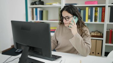 Young beautiful hispanic woman student using computer talking on smartphone at library university