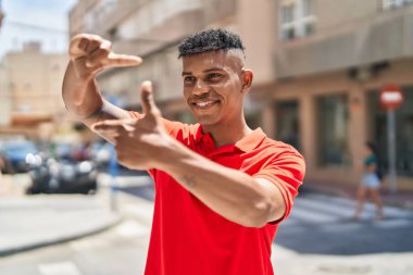Genç Latin adam gülümsüyor. Kendine güveniyor. Sokakta elleriyle fotoğraf hareketi yapıyor.