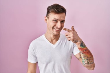 Pembe arka planda duran beyaz bir adam, parmakları burnuna ve yüzüne dönük, neşeli bir şekilde gülümsüyor. güzellik konsepti 