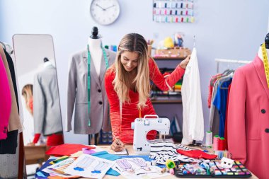 Young blonde woman tailor smiling confident holding t shirt writing on notebook at tailor shop